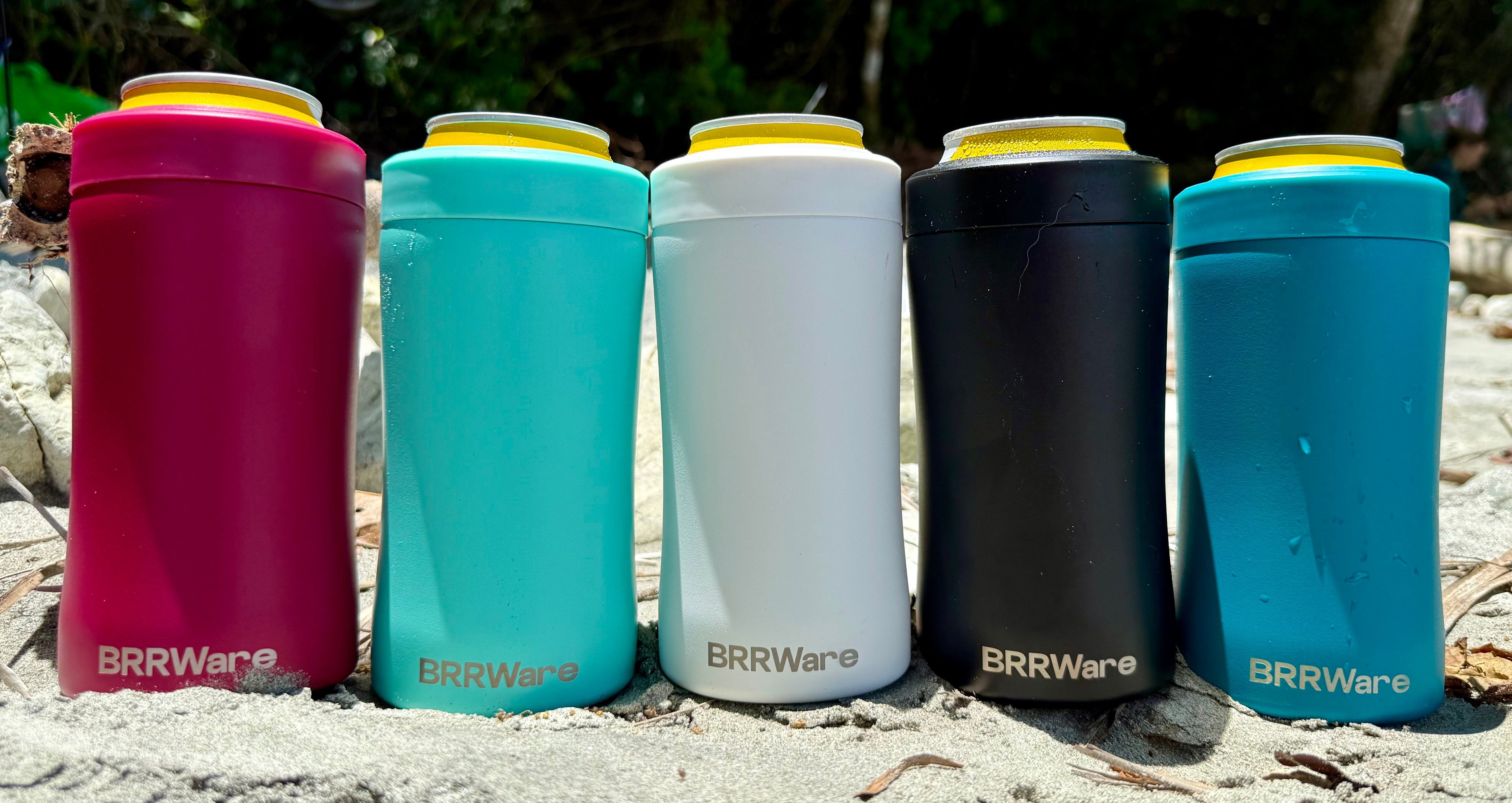 Five BRRRWare insulated drink containers in various colors on a sandy surface with a natural background.